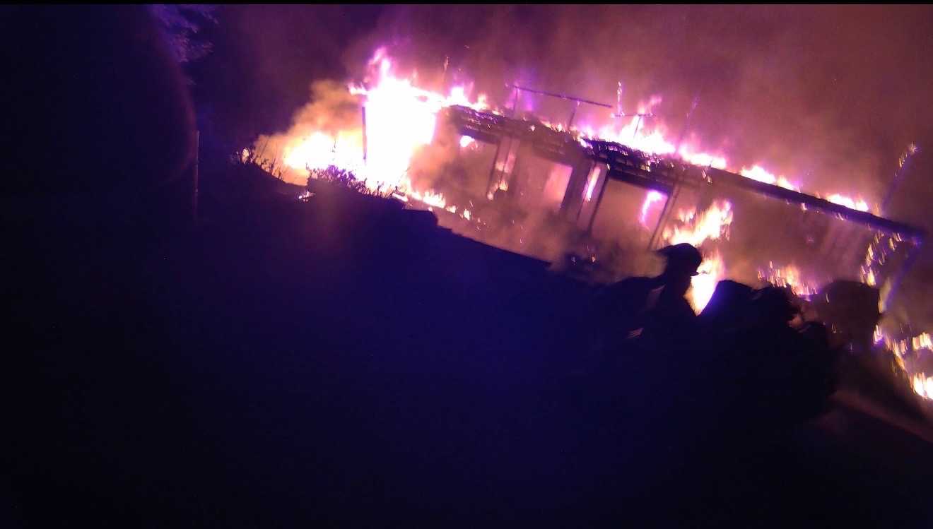 Flames engulf a single-family home during a nighttime residential fire as heavy smoke rises into the air. A firefighter is silhouetted in the foreground while fire spreads across the roof and interior of the structure.